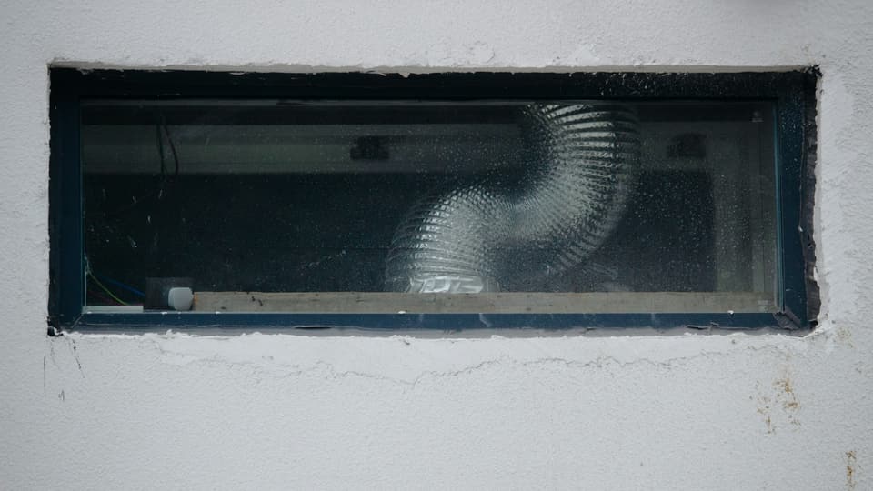 A coil of silver flexible duct seen through a small window in a white wall