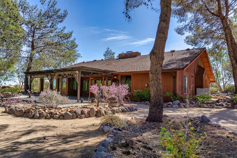 A single-storey wood-sided ranch house with a pergola, rock-edged garden beds and tall pines