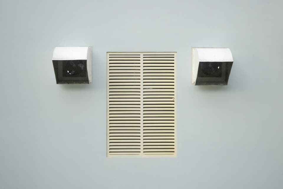 A cream louvered grille on a pale wall between two small exhaust hoods